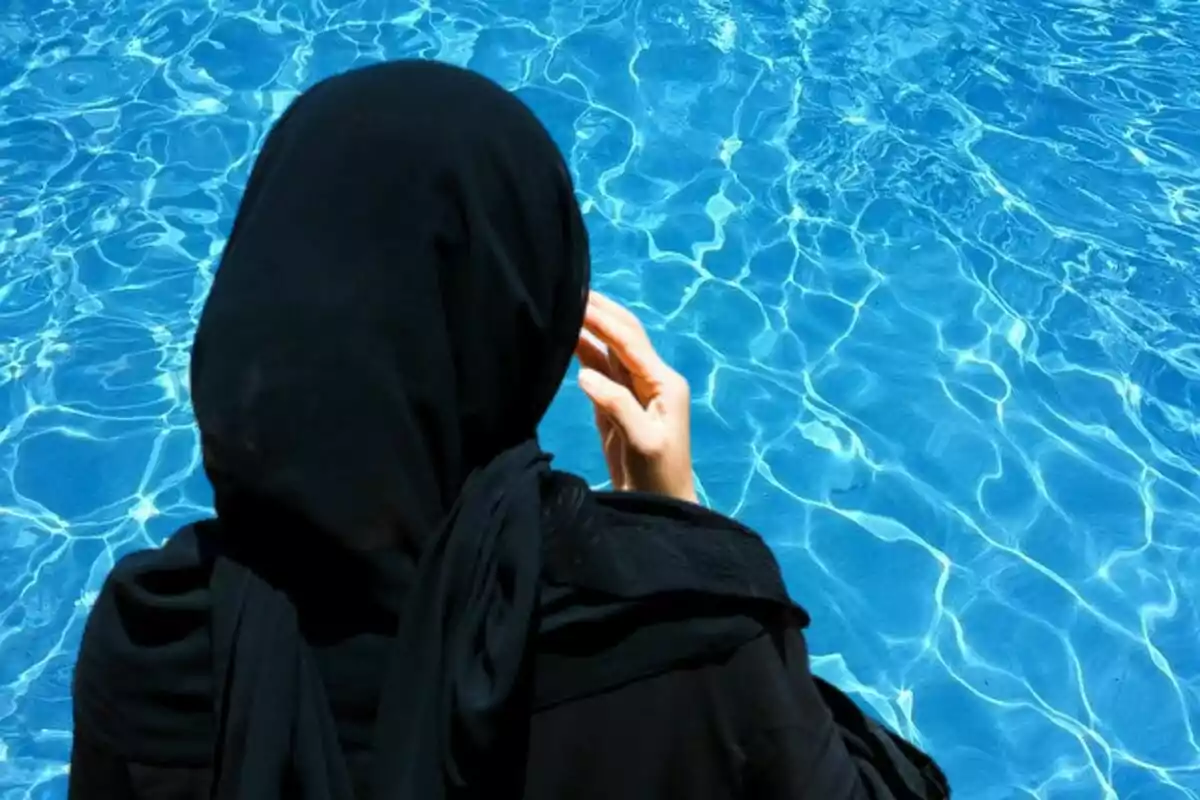 Mujer con hiyab negro de espaldas junto a una piscina de agua azul