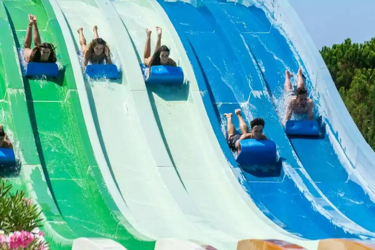 Varias personas deslizándose en colchonetas por toboganes de agua de colores verde y azul en un parque acuático bajo un cielo despejado Varias personas deslizándose en colchonetas por toboganes de agua de colores verde y azul en un parque acuático bajo un cielo despejado