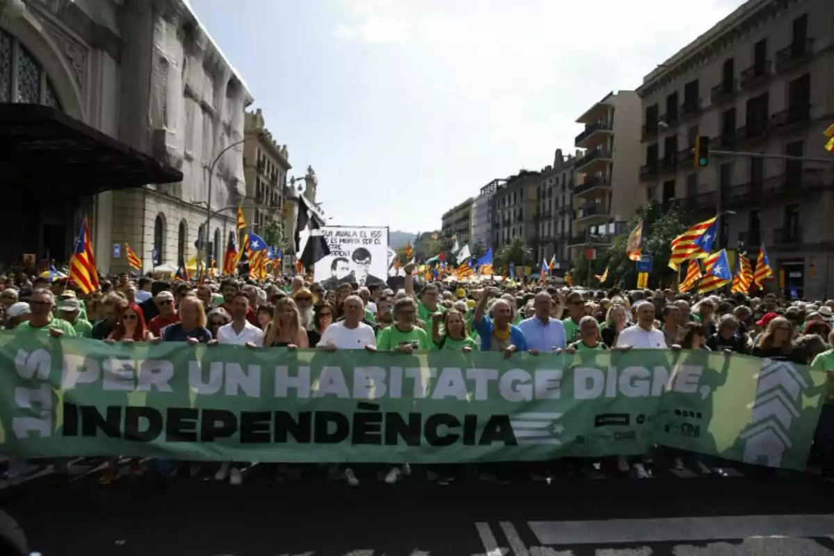 Una multitud de personas marchando en una manifestación con una pancarta verde que dice "PER UN HABITATGE DIGNE, INDEPENDÈNCIA" y banderas catalanas ondeando en el fondo.