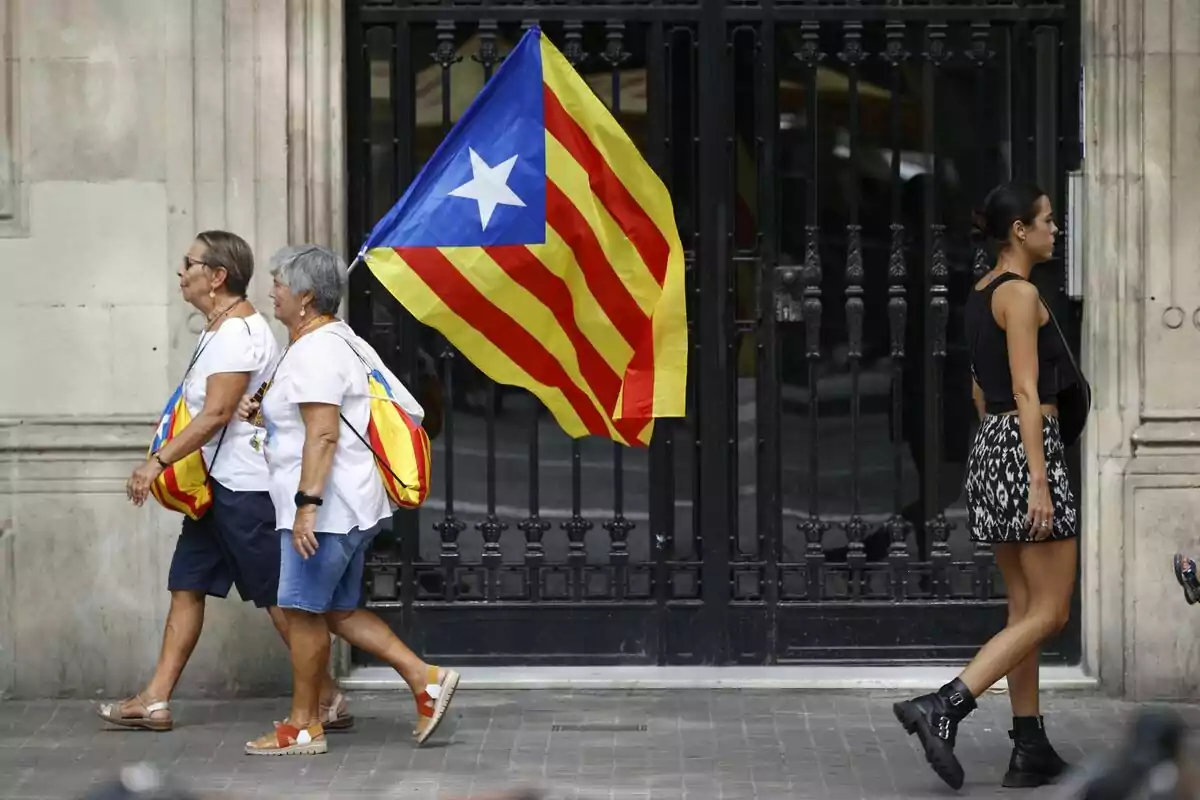 Dos mujeres mayores caminando junto a una bandera estelada mientras una mujer joven pasa por el lado opuesto.