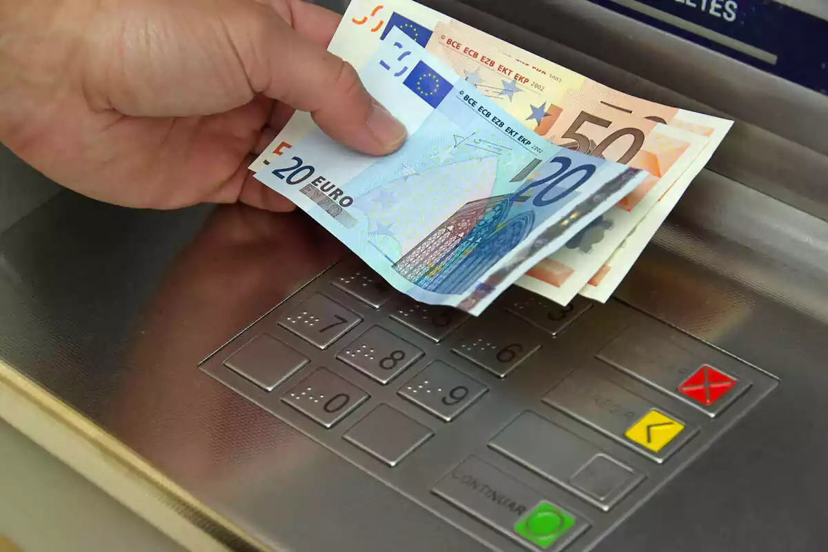 Mano izquierda de una persona sosteniendo un billete de 20 euros y dos billetes de 50 euros, en un cajero automático