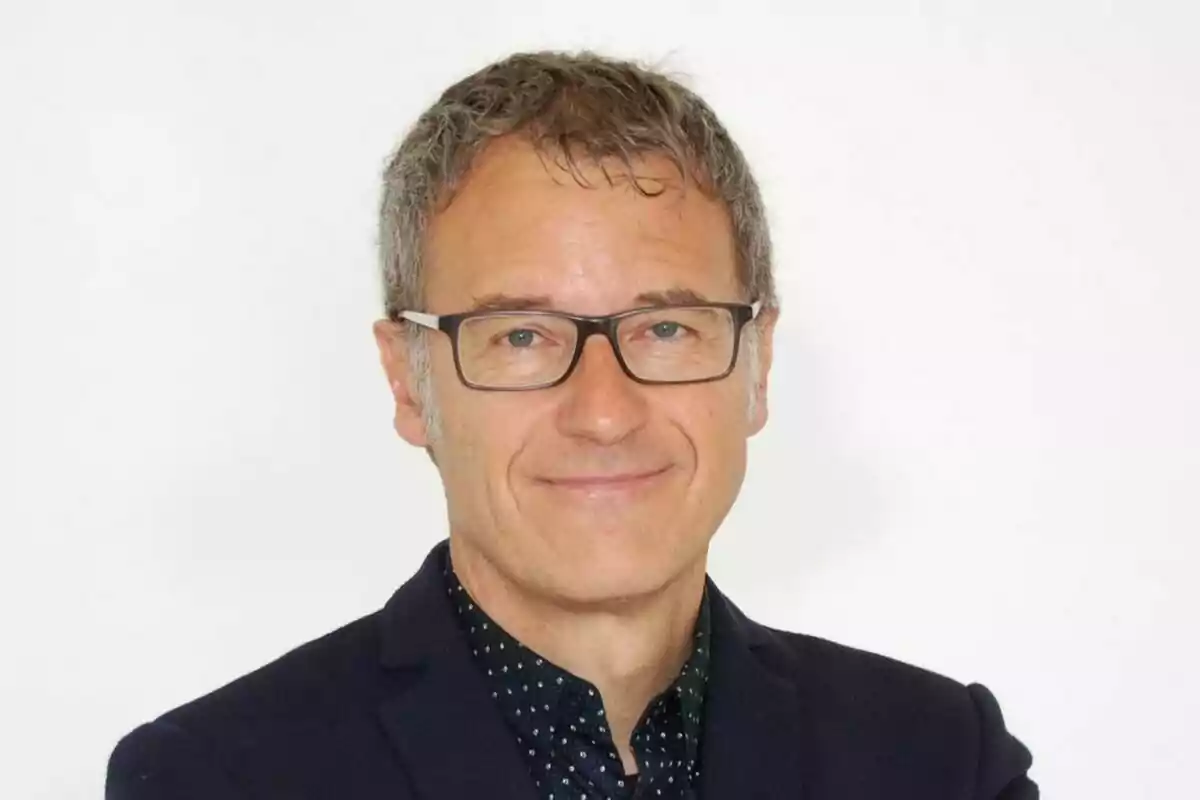 Hombre de cabello corto y canoso con gafas y chaqueta oscura sonriendo frente a un fondo blanco Hombre de cabello corto y canoso con gafas y chaqueta oscura sonriendo frente a un fondo blanco
