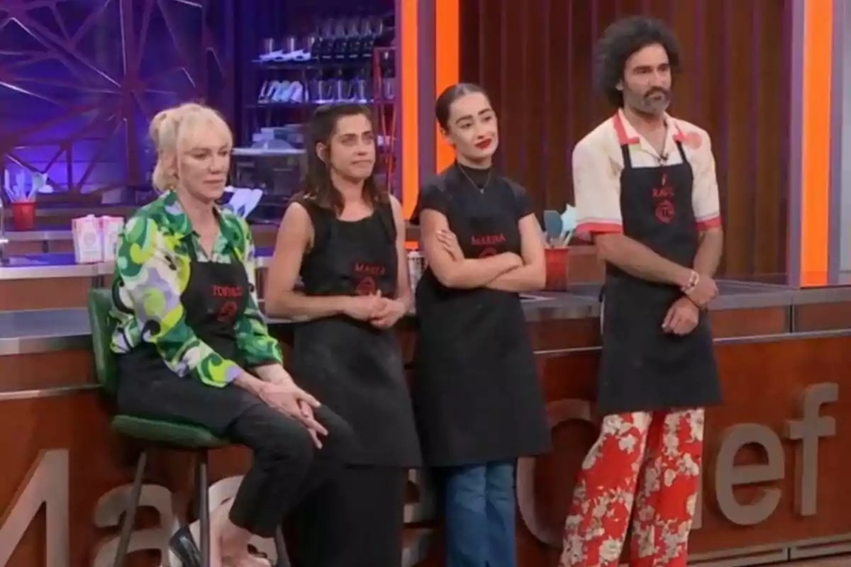 Topacio Fresh, María León, Marina Rivers y Raúl Gómez con delantales negros están de pie en un set de cocina, con una decoración moderna y utensilios de cocina al fondo en MasterChef Celebrity.