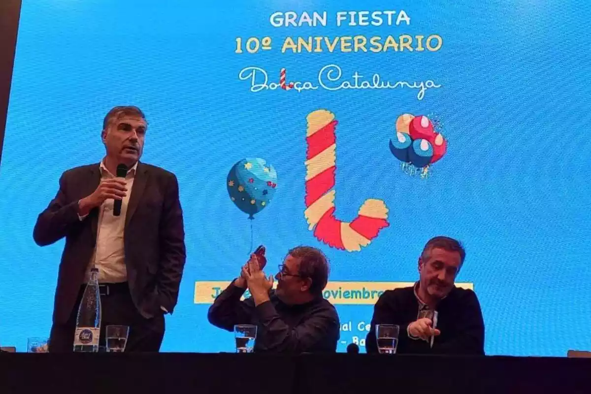 Joan López Alegre de pie y Sergio Fidalgo y Pau Guix sentados y detrás una imagen azul con el logo de Dolça Catalunya