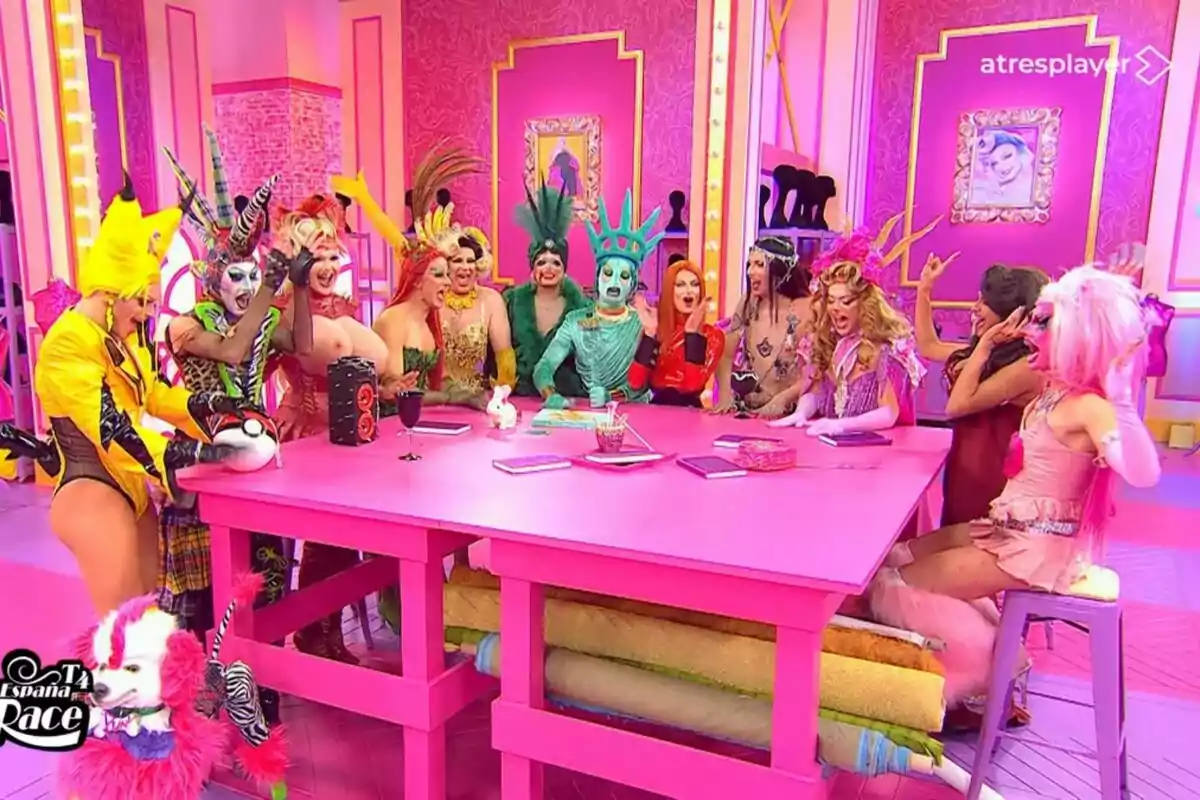 Las reinas con disfraces coloridos y extravagantes se encuentran reunidas alrededor de una mesa rosa en un set de televisión decorado con tonos rosados y detalles dorados en Drag Race España.