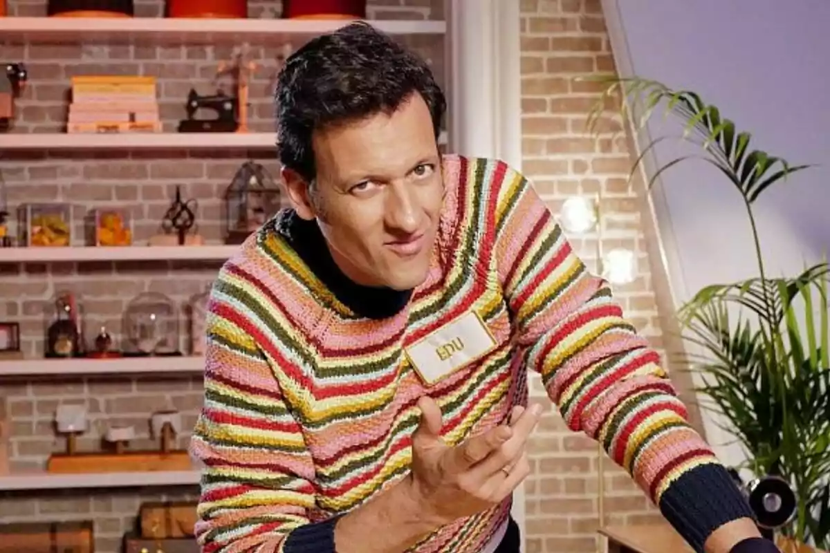 Edu Soto con suéter de rayas coloridas y etiqueta de nombre en un entorno de cocina con fondo de ladrillos y estantes en Maestros de la Costura Celebrity. Edu Soto con suéter de rayas coloridas y etiqueta de nombre en un entorno de cocina con fondo de ladrillos y estantes en Maestros de la Costura Celebrity.