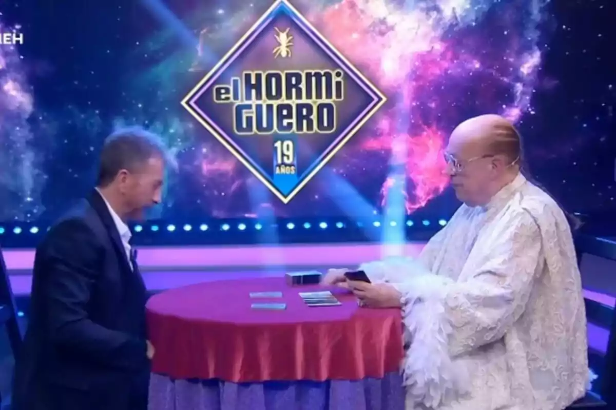 Pablo Motos y Rappel están sentadas en una mesa redonda cubierta con un mantel rojo en un set de televisión con un fondo colorido que muestra el logotipo de "El Hormiguero 19 años".