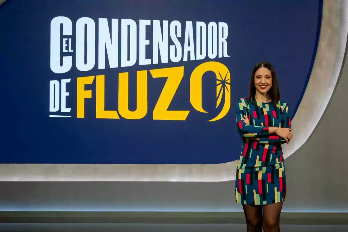 Maya Pixelskaya sonriente con un vestido colorido está de pie frente a un cartel que dice "El Condensador de Fluzo".