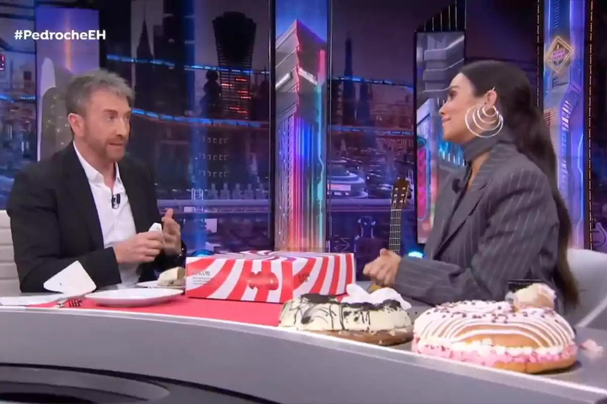 Pablo Motos y Cristina Pedroche conversan en 'El Hormiguero' con un fondo colorido y varios pasteles sobre la mesa.