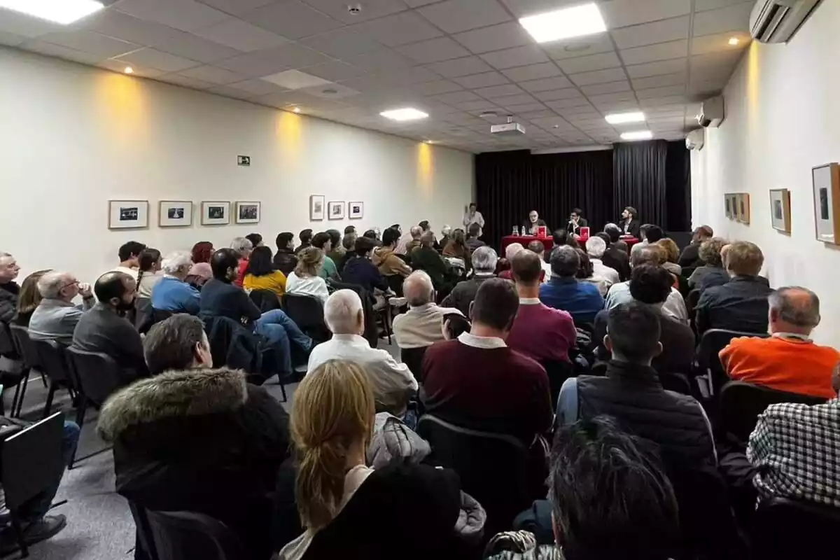 Decenas de personas, sentadas y de espaldas, siguen un acto presentado por tres personas que presiden la sala en el fondo de la imagen