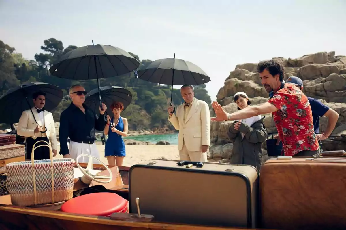 Un grupo de personas encabezado por José Coronado con sombrillas negras está de pie en una playa junto a un bote, mientras un hombre con camisa hawaiana parece estar dando instrucciones en el rodaje de la serie El Problema Final. Un grupo de personas encabezado por José Coronado con sombrillas negras está de pie en una playa junto a un bote, mientras un hombre con camisa hawaiana parece estar dando instrucciones en el rodaje de la serie El Problema Final.