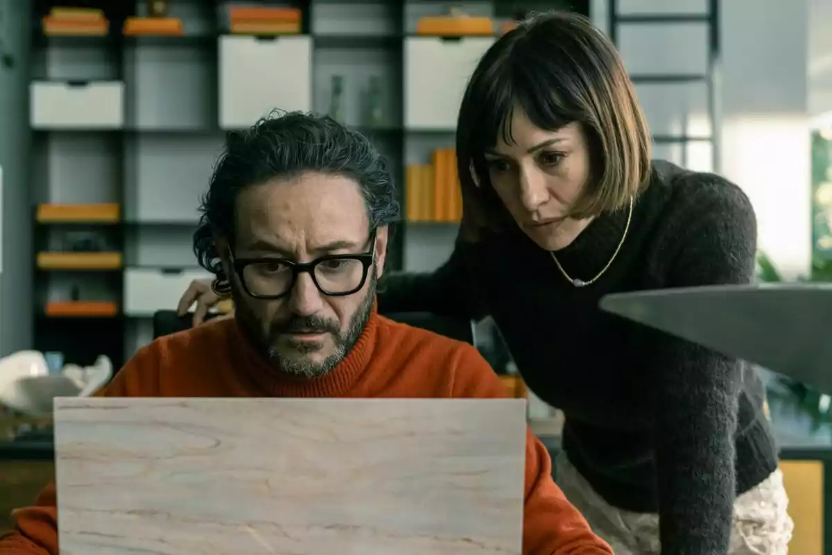 Carlos Santos con gafas y suéter naranja mira preocupado la pantalla de una laptop mientras una mujer de cabello corto y suéter oscuro lo observa atentamente desde su lado en una oficina moderna en El Refugio Atómico. Carlos Santos con gafas y suéter naranja mira preocupado la pantalla de una laptop mientras una mujer de cabello corto y suéter oscuro lo observa atentamente desde su lado en una oficina moderna en El Refugio Atómico.