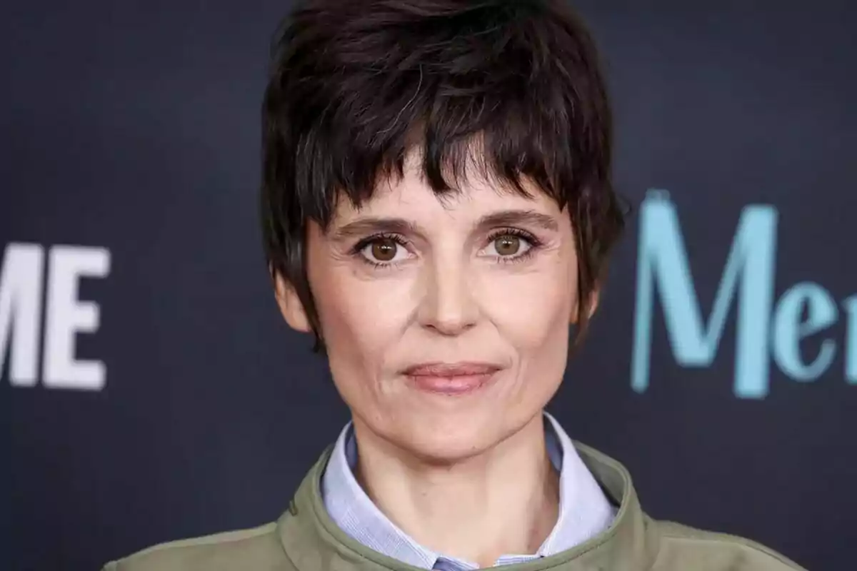 Fotografía en primer plano de Elena Anaya con cabello corto y oscuro, vistiendo una chaqueta verde y camisa azul, posando frente a un fondo oscuro con texto parcialmente visible.