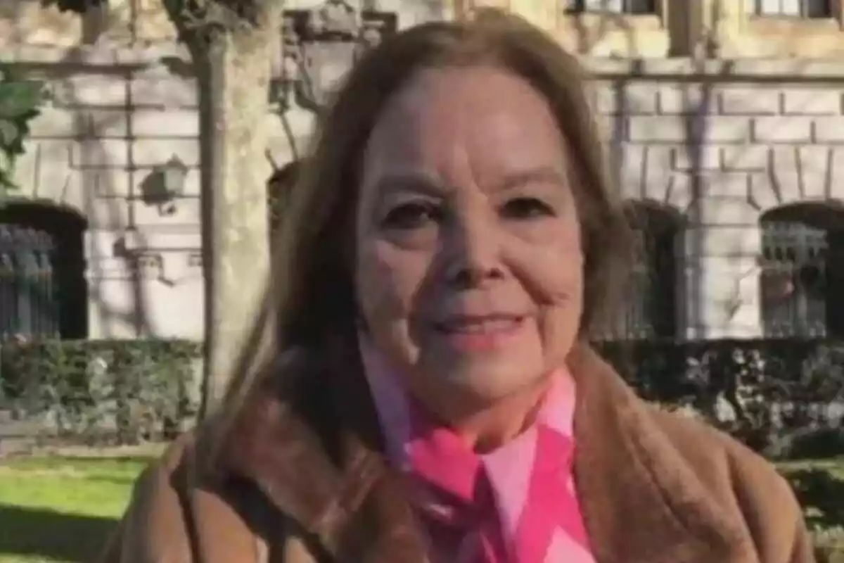 Elisa Montés de cabello castaño claro y abrigo marrón está sonriendo al aire libre con un edificio de piedra y árboles al fondo.