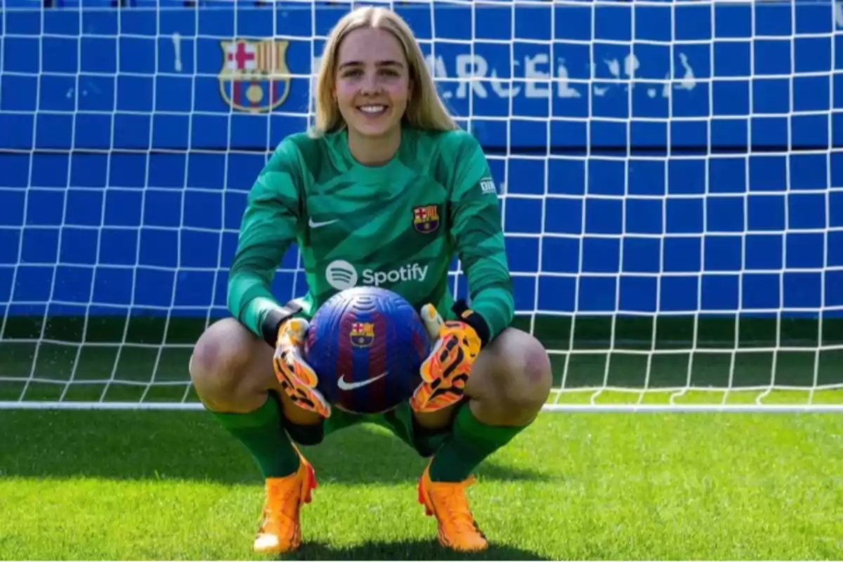 Portera del FC Barcelona femenino sonríe frente a la portería mientras sostiene un balón con ambas manos y viste uniforme verde con guantes y botas naranjas Portera del FC Barcelona femenino sonríe frente a la portería mientras sostiene un balón con ambas manos y viste uniforme verde con guantes y botas naranjas
