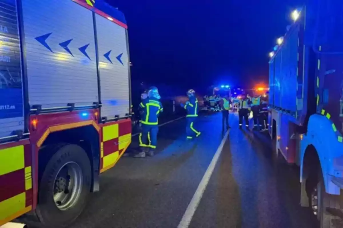 Camiones de bomberos y personal de emergencia trabajando en una carretera por la noche con luces azules y amarillas iluminando la escena.