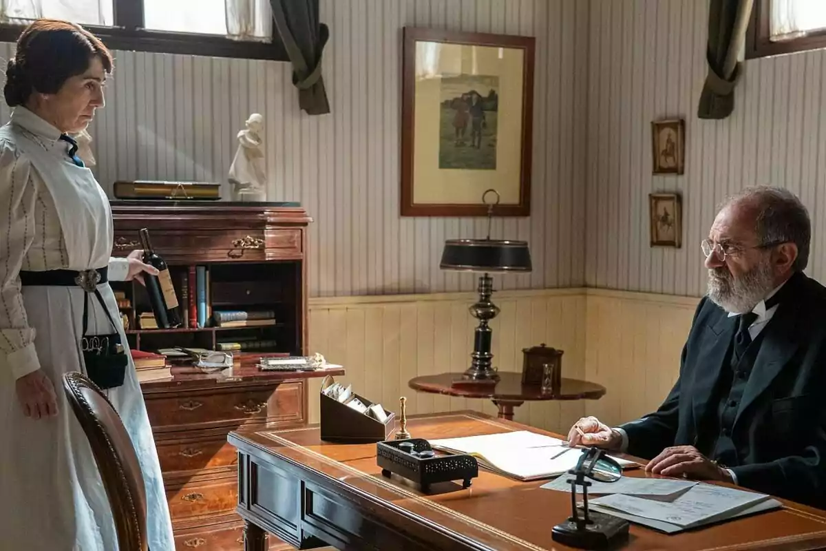 Arantxa Aranguren con uniforme de servicio sostiene una botella de vino mientras habla con Joaquín Climent mayor sentado en un escritorio en una oficina decorada con muebles de madera y cuadros en las paredes en La Promesa. Arantxa Aranguren con uniforme de servicio sostiene una botella de vino mientras habla con Joaquín Climent mayor sentado en un escritorio en una oficina decorada con muebles de madera y cuadros en las paredes en La Promesa.