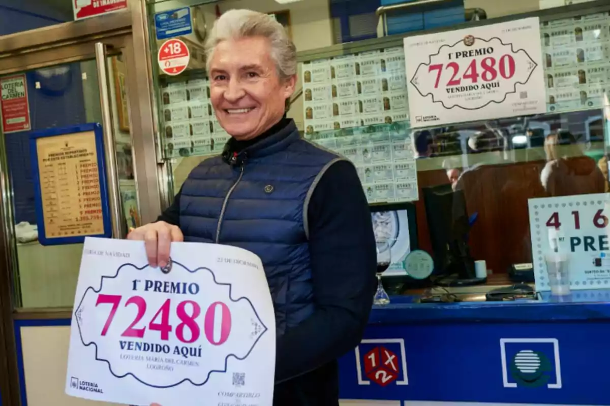 Un hombre sonriente sostiene un cartel que anuncia el primer premio de la lotería con el número 72480 en una administración de lotería.