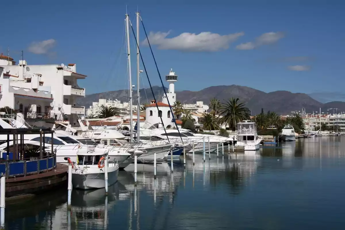 Puerto deportivo con yates atracados, edificios blancos y un faro al fondo, con montañas y cielo azul claro. Puerto deportivo con yates atracados, edificios blancos y un faro al fondo, con montañas y cielo azul claro.