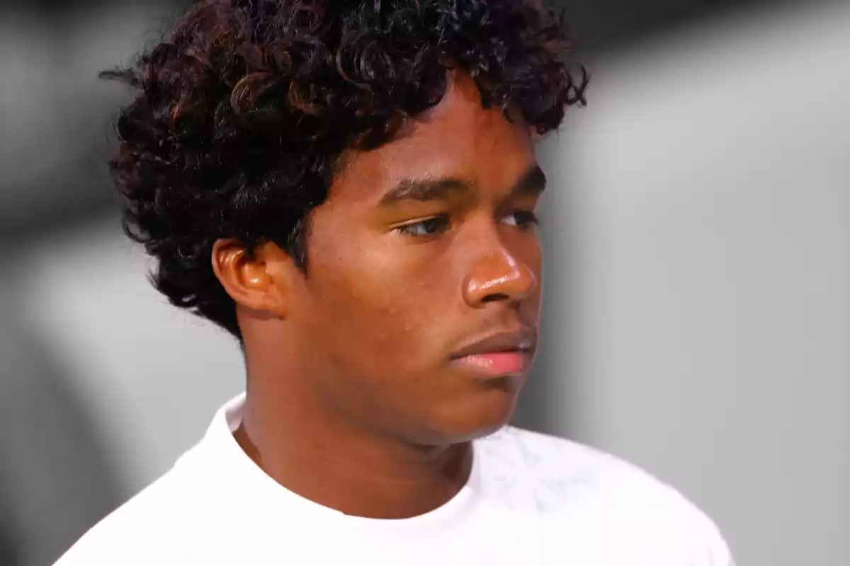 Joven con cabello rizado y expresión seria, vistiendo una camiseta blanca.