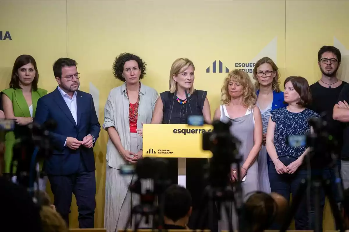 Un grupo de personas de pie frente a un podio amarillo con el logotipo de Esquerra Republicana, mientras varias cámaras los graban.