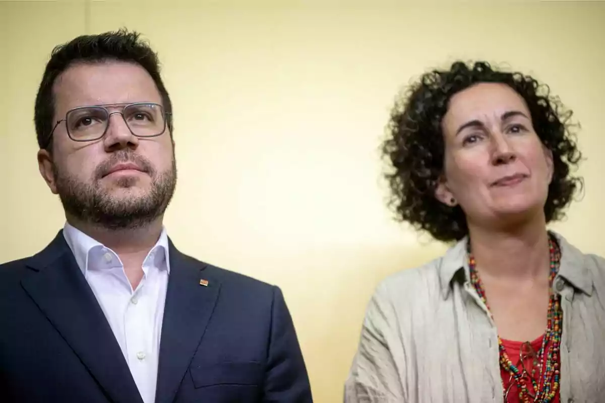 Dos personas de pie, una con traje y gafas y la otra con camisa clara y collar colorido, sobre un fondo amarillo.