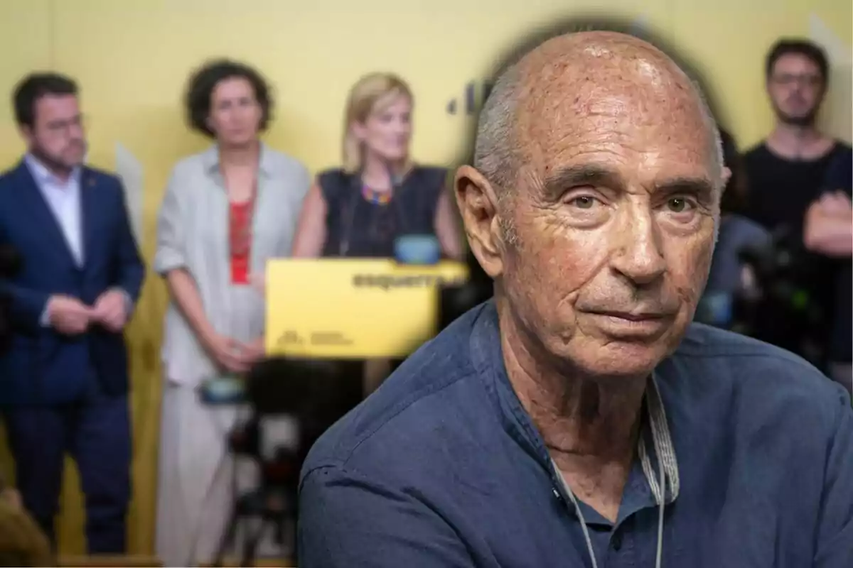 Un hombre mayor con camisa azul aparece en primer plano, mientras que en el fondo se ven varias personas de pie frente a un podio con un cartel amarillo.