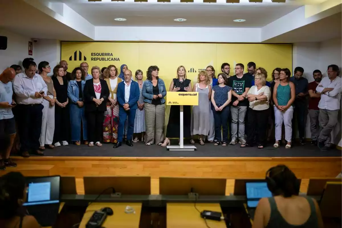 Un grupo de personas de pie en un escenario con un fondo amarillo y un podio en el centro.