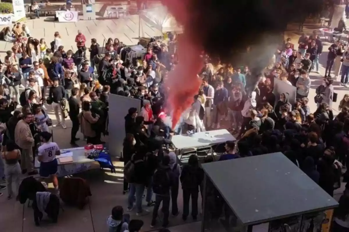 Un grupo numeroso de personas se reúne alrededor de una mesa mientras una columna de humo rojo y negro se eleva en el centro del evento al aire libre.