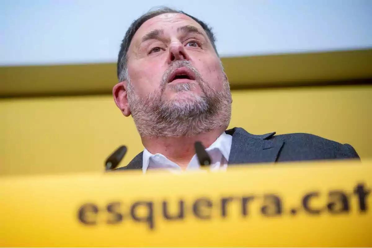 Hombre hablando en un podio con el logo de "esquerra.cat" en un fondo amarillo.