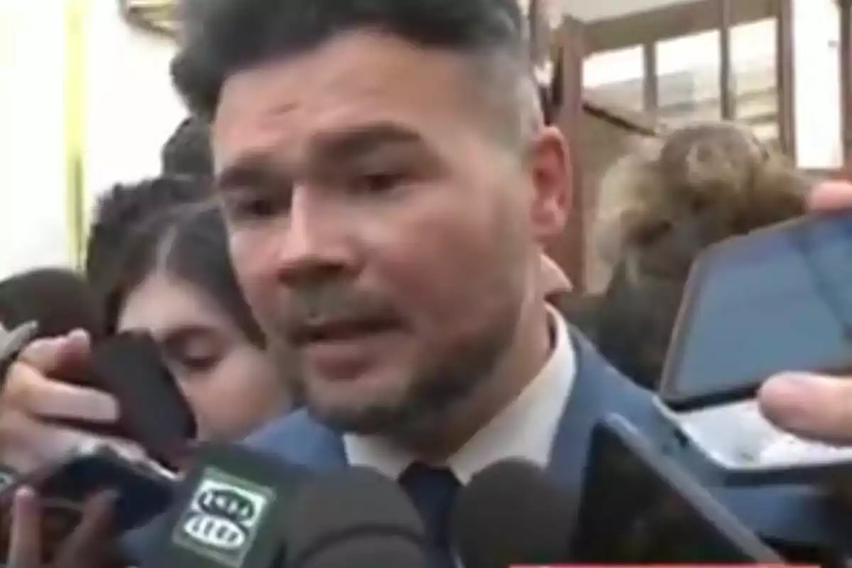 Hombre con barba y traje azul hablando con la prensa rodeado de micrófonos y teléfonos móviles