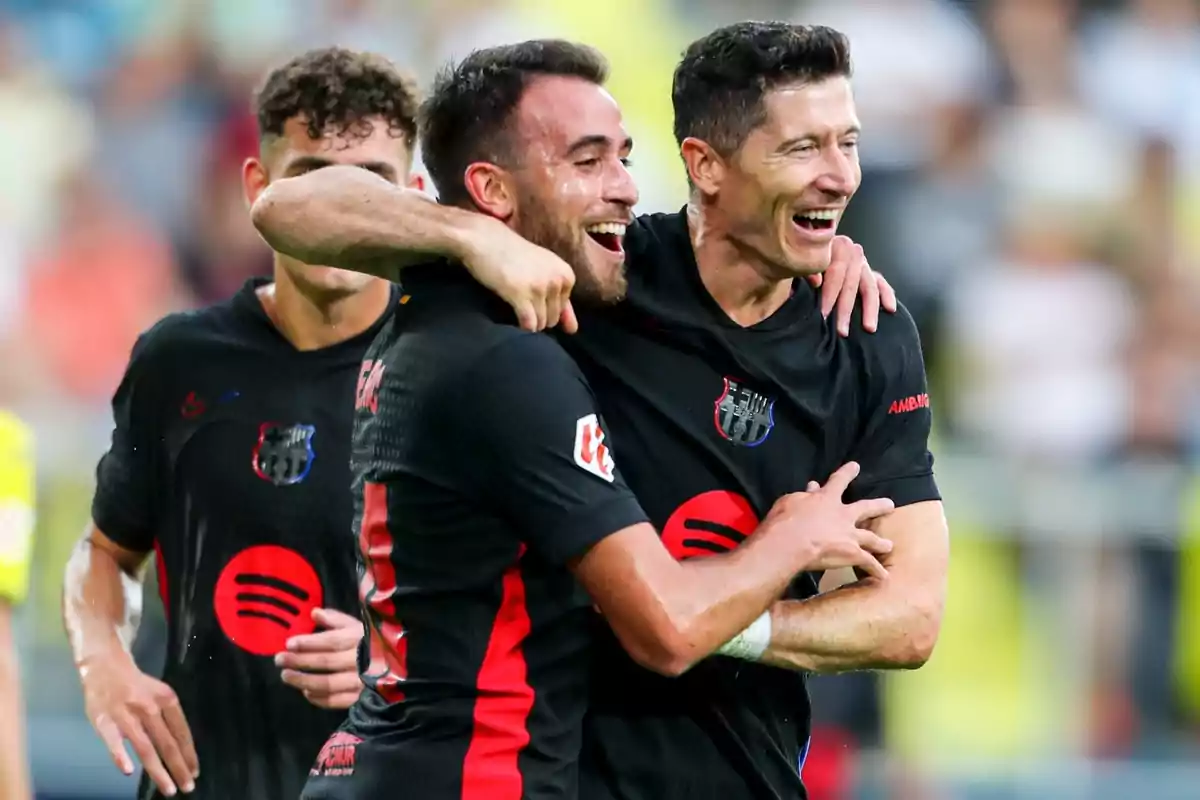 Jugadores de fútbol celebrando un gol durante un partido, vistiendo uniformes negros con detalles rojos y el logo de un patrocinador en el pecho.