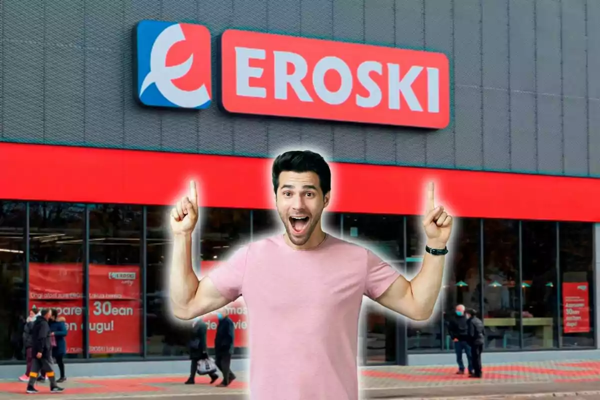 Un hombre sonriente con camiseta rosa señala hacia arriba con ambos dedos índices frente a una tienda Eroski.