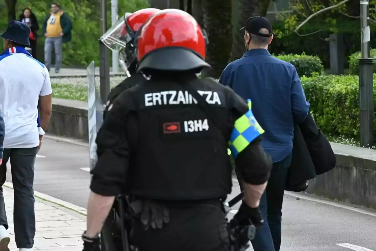 Agente de la Ertzaintza con casco rojo y uniforme negro caminando por la calle junto a otras personas. Agente de la Ertzaintza con casco rojo y uniforme negro caminando por la calle junto a otras personas.