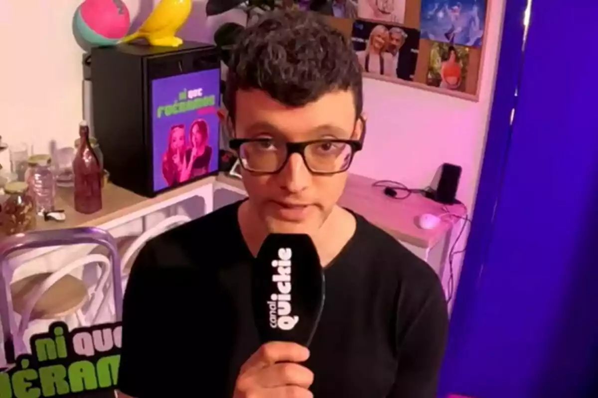 César Toral (Escaleto) con gafas sostiene un micrófono de "canal Quickie" en una habitación de Ni que fuéramos decorada con varios objetos y fotos en la pared.