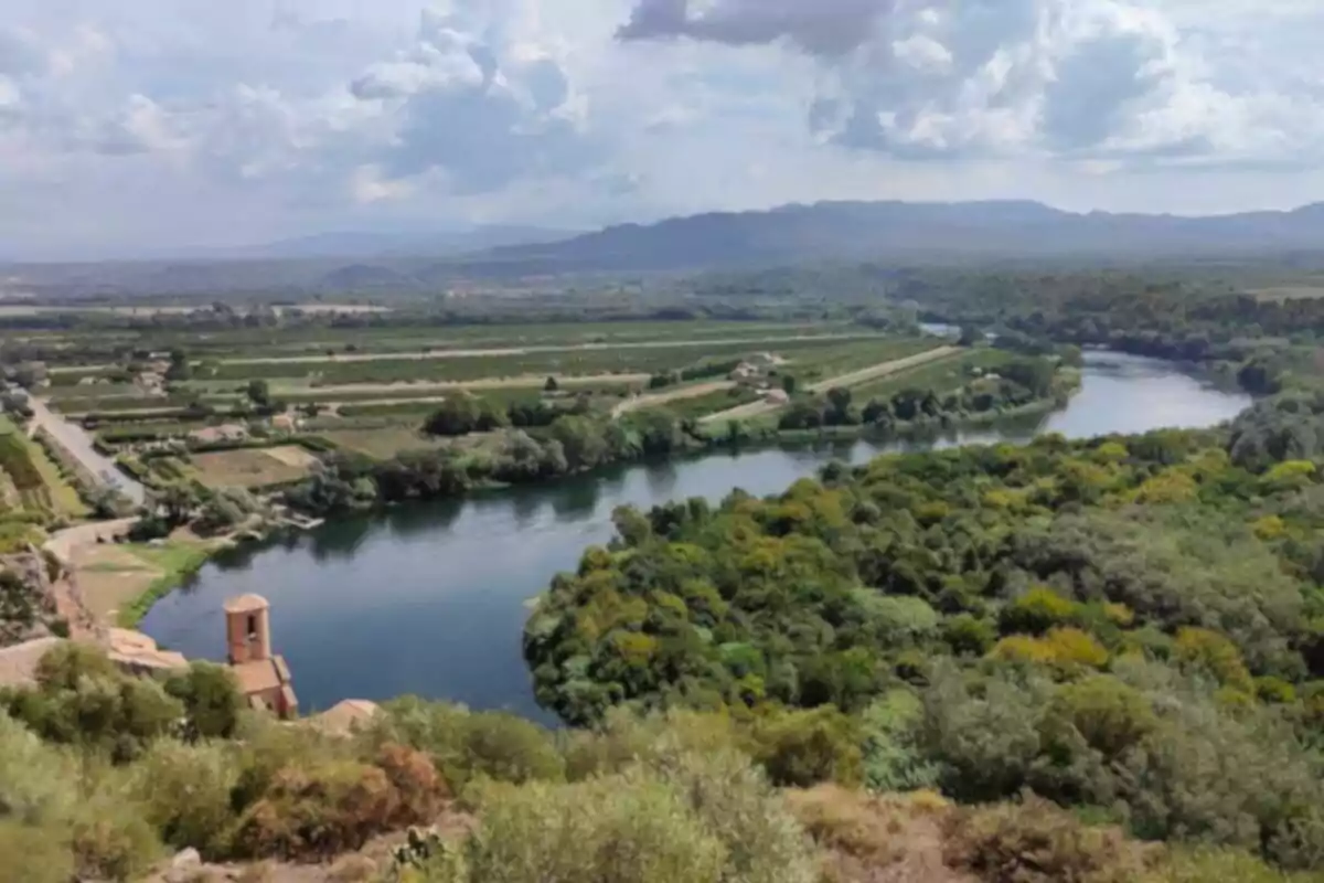 Vista panorámica de un río serpenteante rodeado de vegetación y campos cultivados con montañas al fondo bajo un cielo parcialmente nublado. Vista panorámica de un río serpenteante rodeado de vegetación y campos cultivados con montañas al fondo bajo un cielo parcialmente nublado.