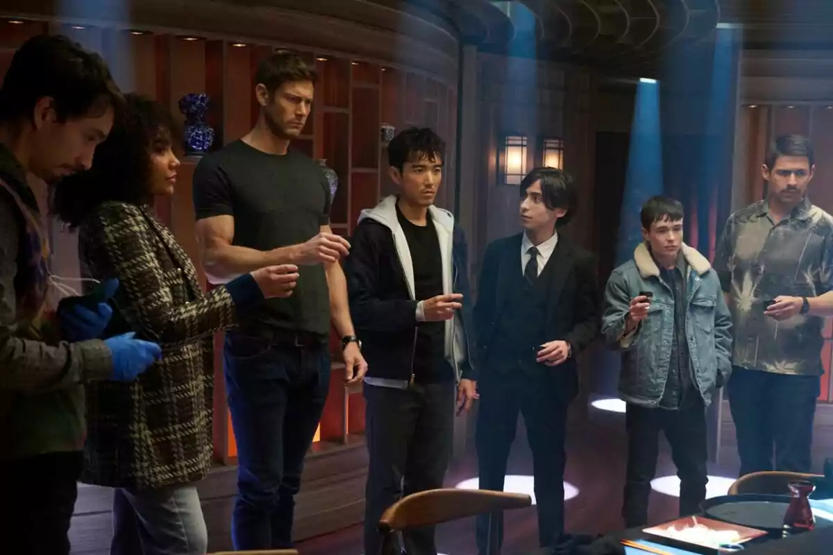 Un grupo de personas de pie en una habitación con iluminación tenue, algunas sosteniendo pequeños objetos en sus manos en una escena de la última temporada de la serie The Umbrella Academy