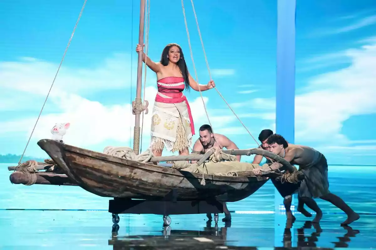 Esperansa Grasia disfrazada de Moana está de pie en una balsa mientras tres hombres la empujan en un escenario con fondo de mar y cielo azul en Tu Cara Me Suena