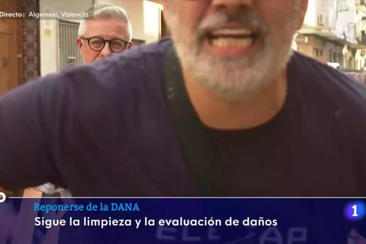 Un hombre con barba aparece en primer plano mientras otro hombre con gafas está al fondo en una calle de Algemesí, Valencia, con un texto en pantalla que menciona la limpieza y evaluación de daños tras la DANA en el Telediario. Un hombre con barba aparece en primer plano mientras otro hombre con gafas está al fondo en una calle de Algemesí, Valencia, con un texto en pantalla que menciona la limpieza y evaluación de daños tras la DANA en el Telediario.