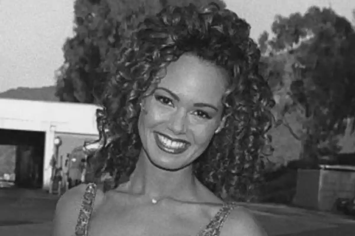 Fotografía en blanco y negro de Esta TerBlanche con cabello rizado y sonrisa amplia, posando al aire libre con árboles y una estructura en el fondo.