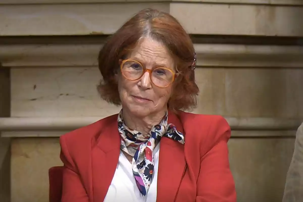 Una mujer mayor con gafas naranjas y cabello castaño, vestida con un saco rojo y una bufanda estampada, está sentada frente a un fondo de piedra.