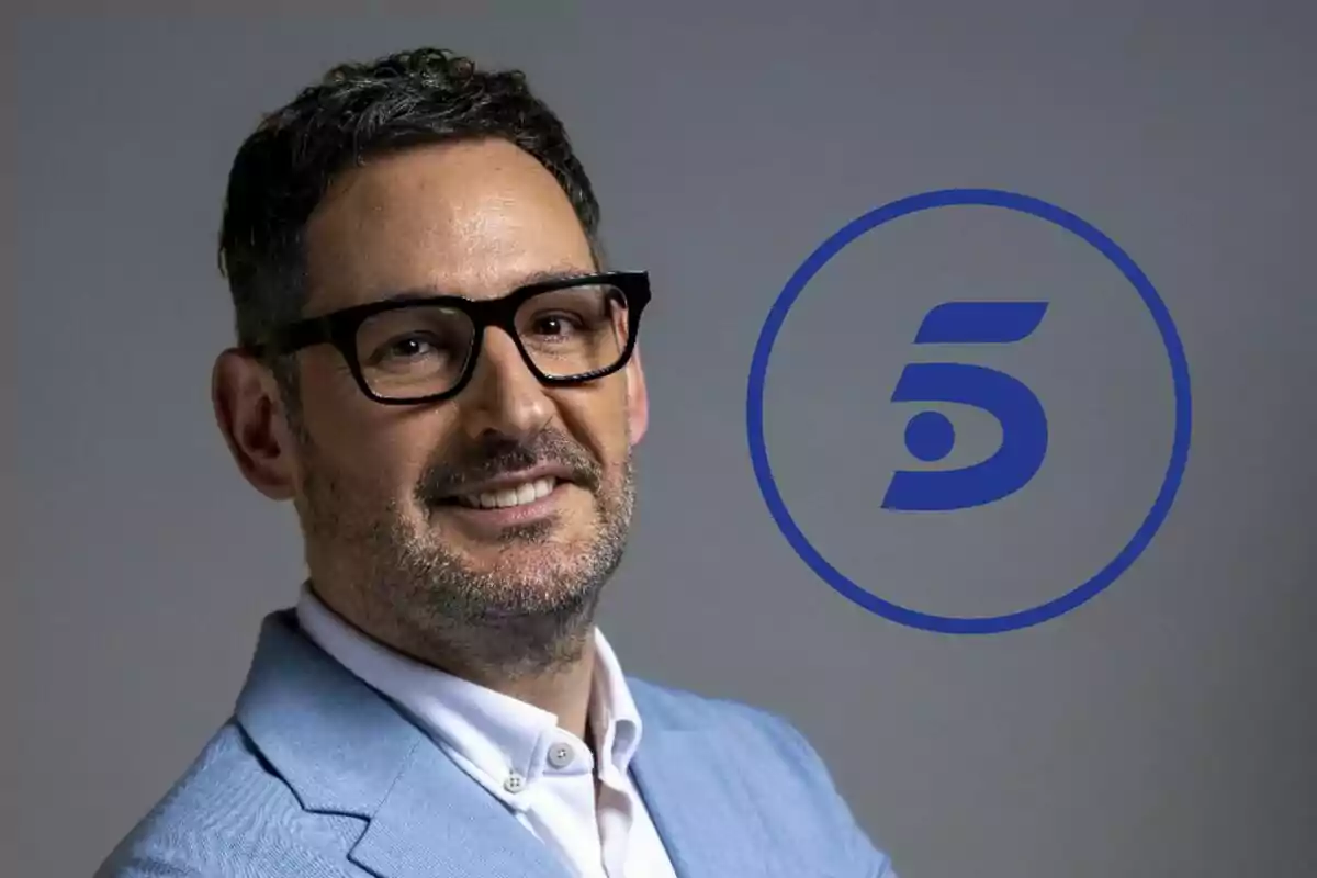 Eugeni Alemany con gafas y barba sonríe vistiendo un saco azul claro junto al logotipo azul de Telecinco sobre fondo gris