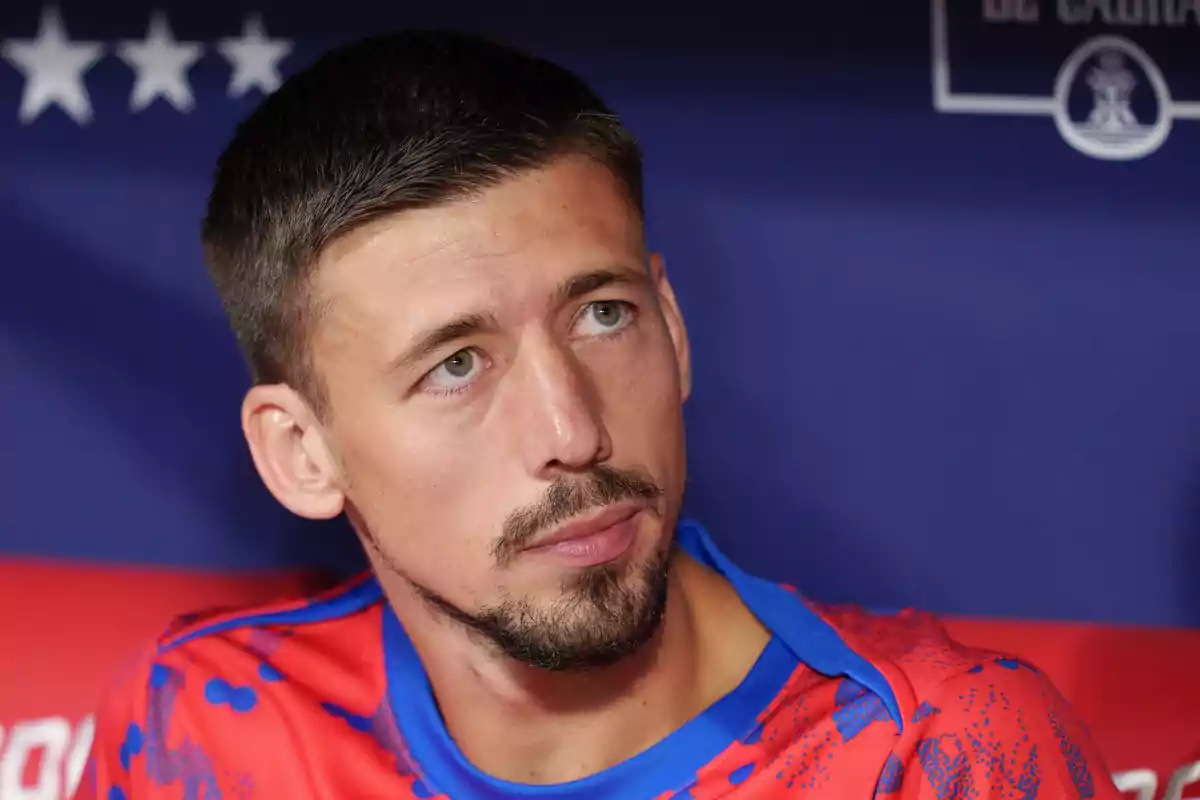 Un hombre con barba y bigote, vistiendo una camiseta roja con detalles azules, mira hacia un lado con una expresión seria. Un hombre con barba y bigote, vistiendo una camiseta roja con detalles azules, mira hacia un lado con una expresión seria.
