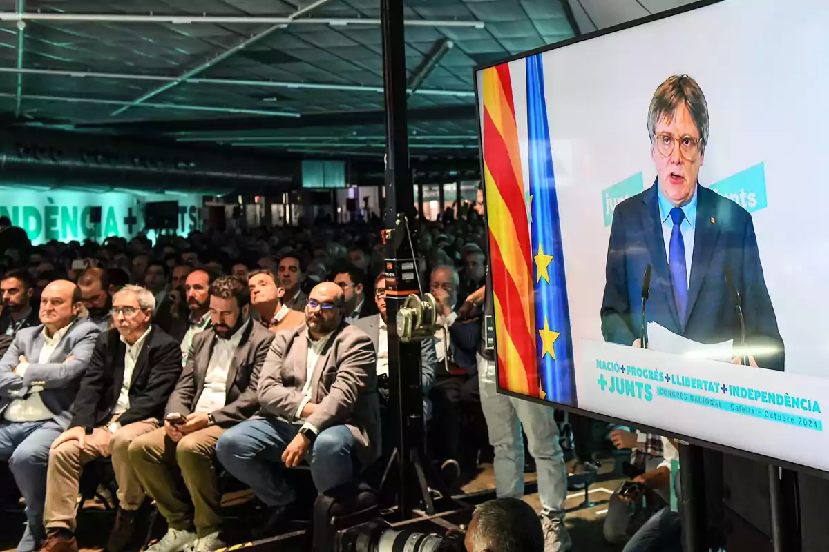 Una multitud de personas sentadas en un evento mientras observan una pantalla grande que muestra a un orador con banderas detrás de él.