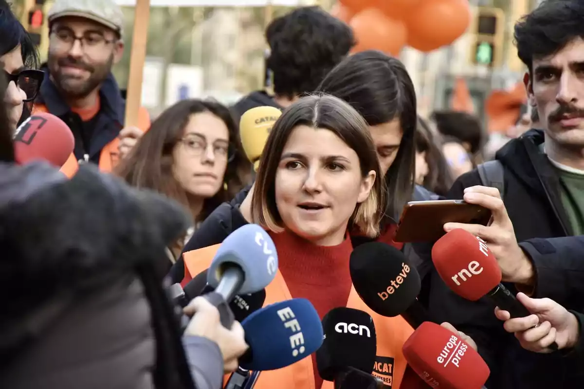 Una mujer con chaleco naranja habla ante varios micrófonos de diferentes medios de comunicación rodeada de personas en un evento al aire libre.