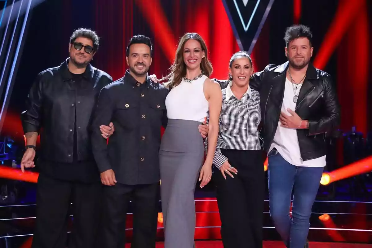 Eva González junto a Pablo López, Antonio Orozco, Luis Fonsi y Malú posan juntas en el plató de La Voz iluminado con luces rojas y negras, con un fondo que incluye un logotipo en forma de "V".