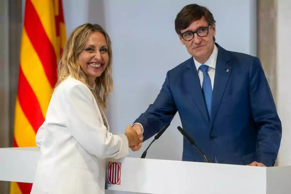 Eva Menor y Salvador Illa estrechándose la mano frente a un podio con una bandera catalana de fondo.