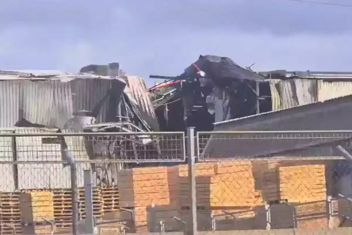 Edificio industrial con daños visibles en el techo y paredes rodeado de una cerca de alambre y pilas de madera en el exterior.