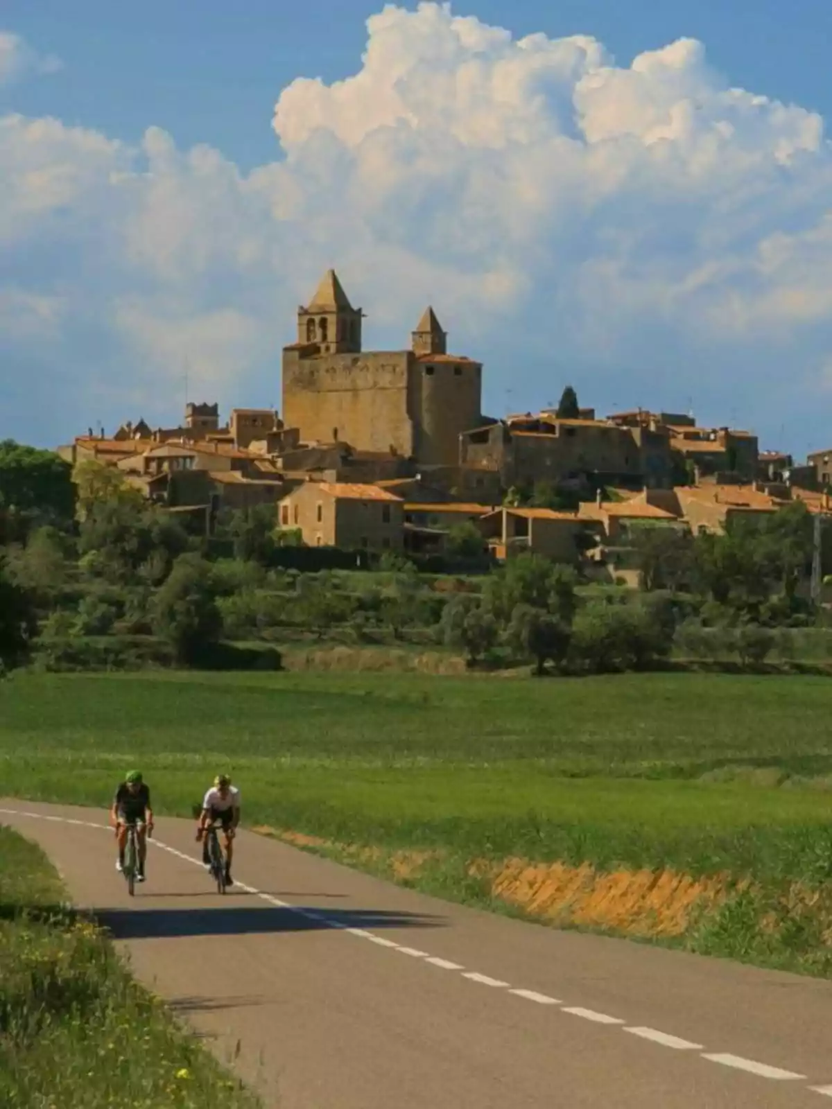 Dos ciclistas recorren una carretera rural con un pintoresco pueblo medieval y un cielo parcialmente nublado al fondo. Dos ciclistas recorren una carretera rural con un pintoresco pueblo medieval y un cielo parcialmente nublado al fondo.