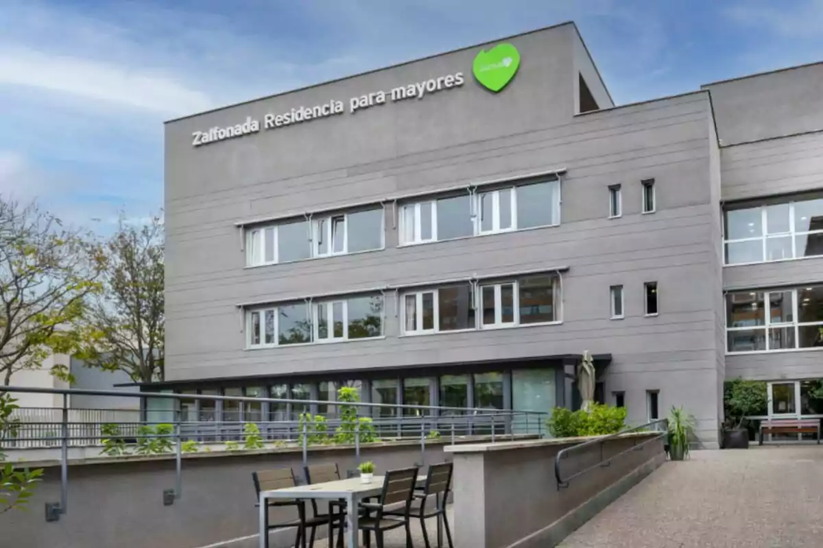 Edificio moderno de tres pisos con letrero que dice Zalfonada Residencia para mayores y una terraza con mesas y sillas en primer plano Edificio moderno de tres pisos con letrero que dice Zalfonada Residencia para mayores y una terraza con mesas y sillas en primer plano
